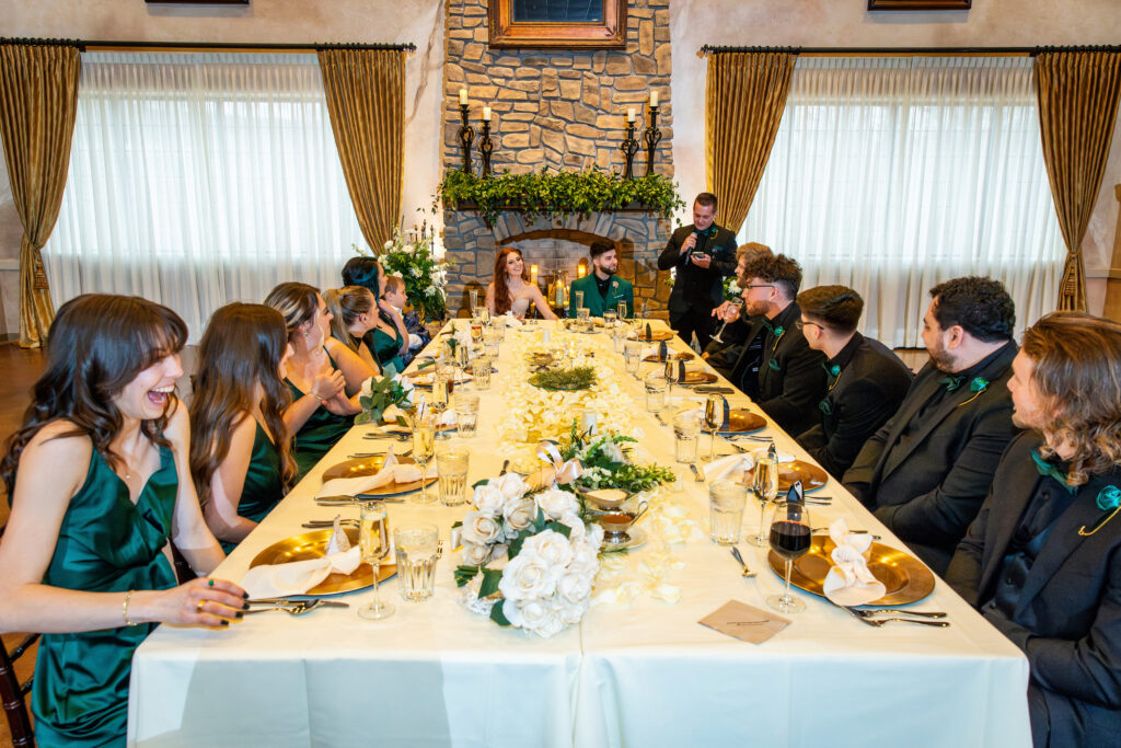 Wedding party seated at a long table, elegantly dressed, with floral centerpieces and draped windows in the background.