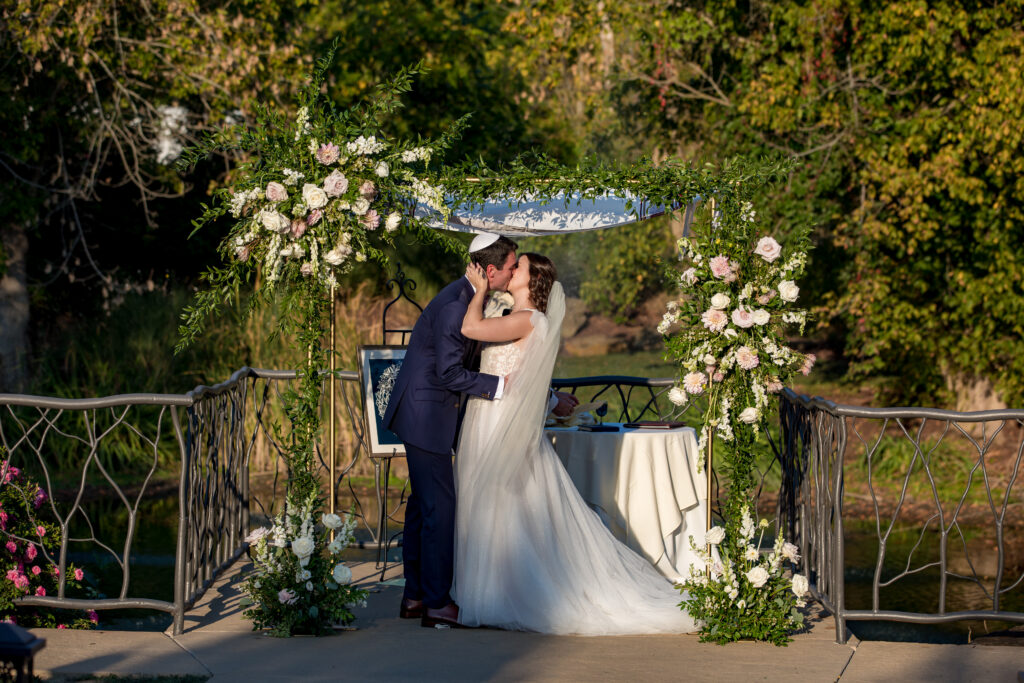 A bride and groom kiss under a floral wedding arch outdoors, surrounded by greenery.