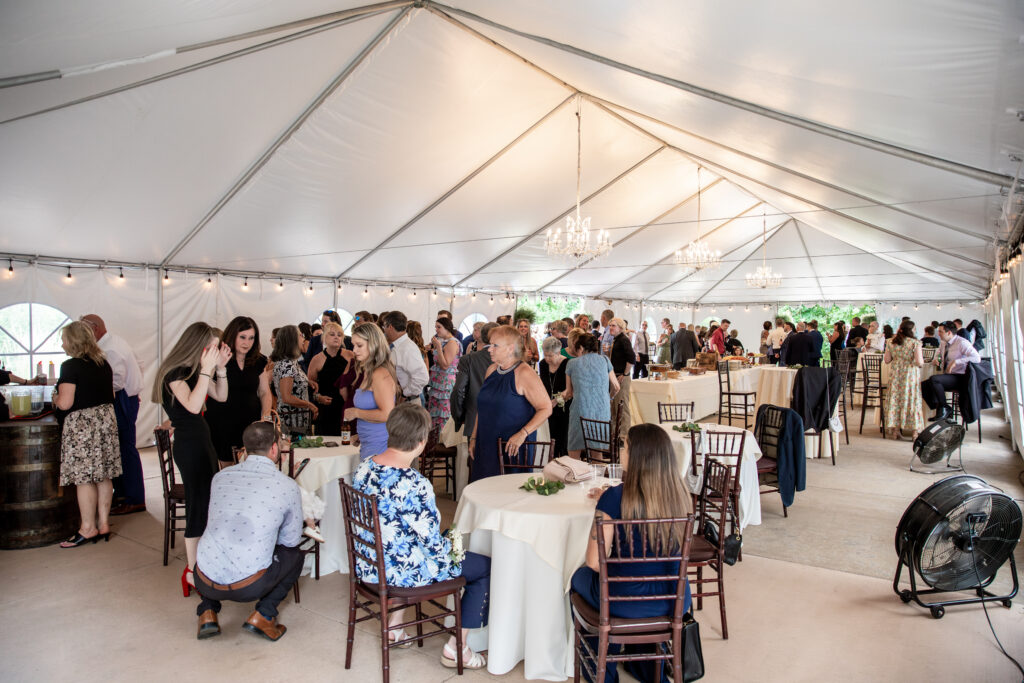 People mingling under a large white tent at a formal event, with tables, chairs, and chandeliers visible.