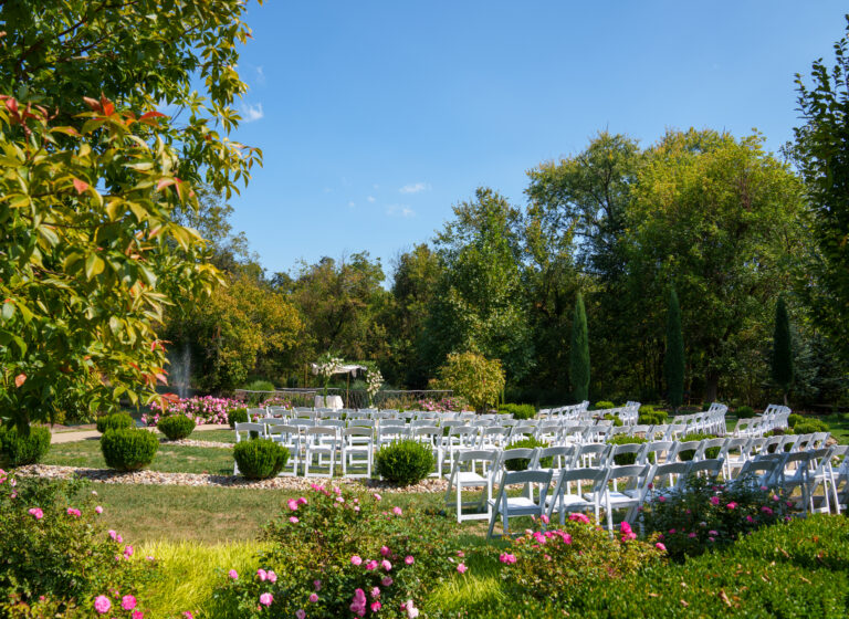 Outdoor wedding setup with white chairs surrounded by greenery and flowers under a clear blue sky.