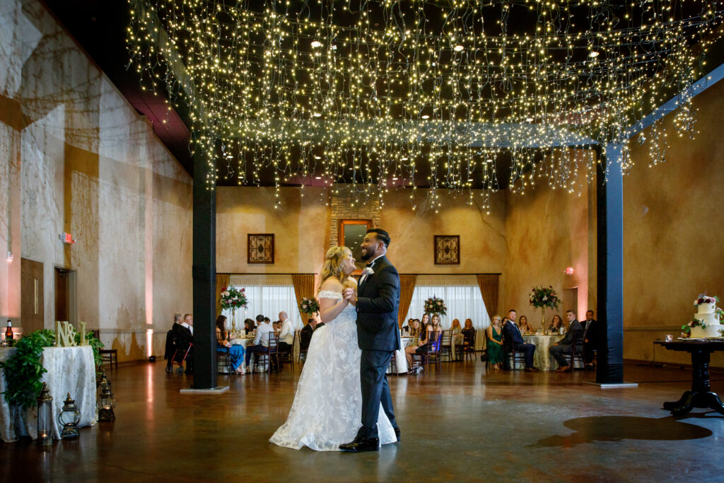 A couple dances under string lights in a warmly lit reception hall, surrounded by seated guests.