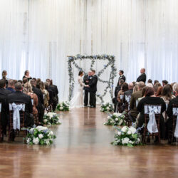 Wedding ceremony with bride and groom under a floral arch, guests seated in rows on either side.