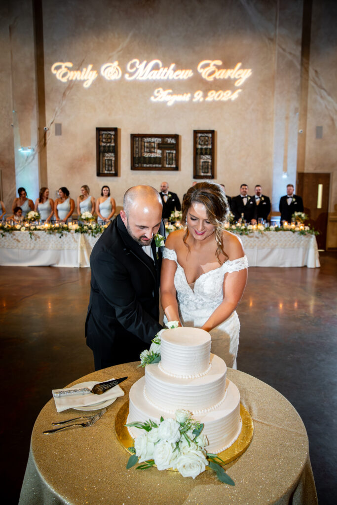 A couple cuts their wedding cake in front of a decorated reception table with their names and date projected above.