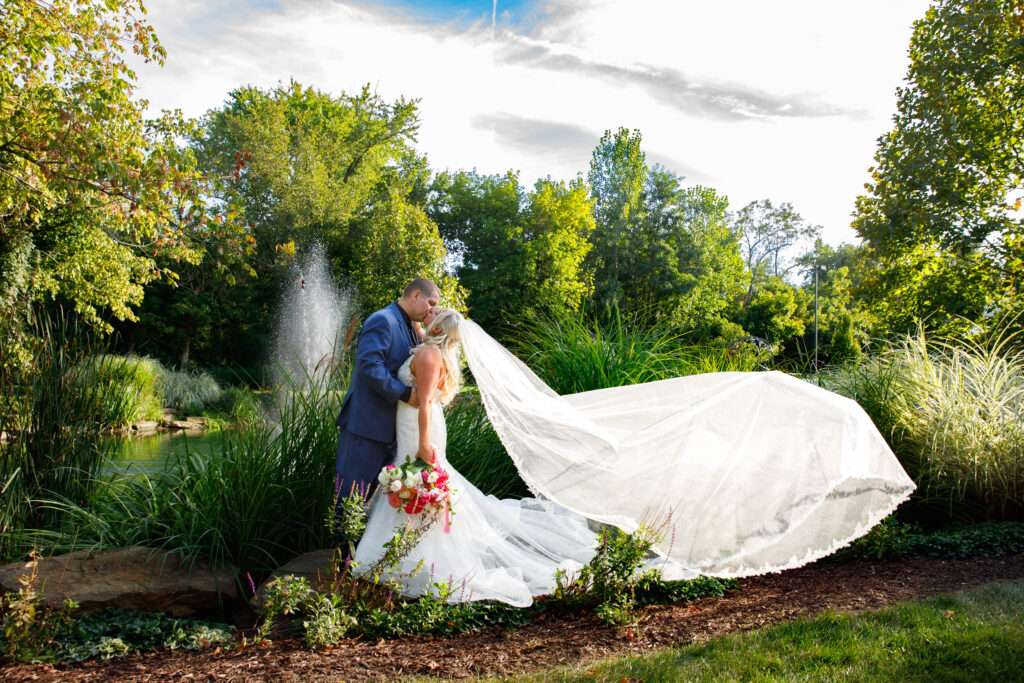 A bride and groom share a kiss near a pond, with her veil flowing and greenery surrounding them.