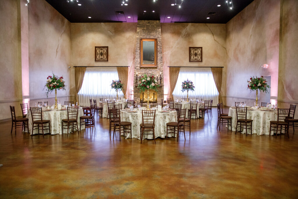 Elegant banquet hall with round tables, floral centerpieces, and a stone fireplace centered on the back wall.