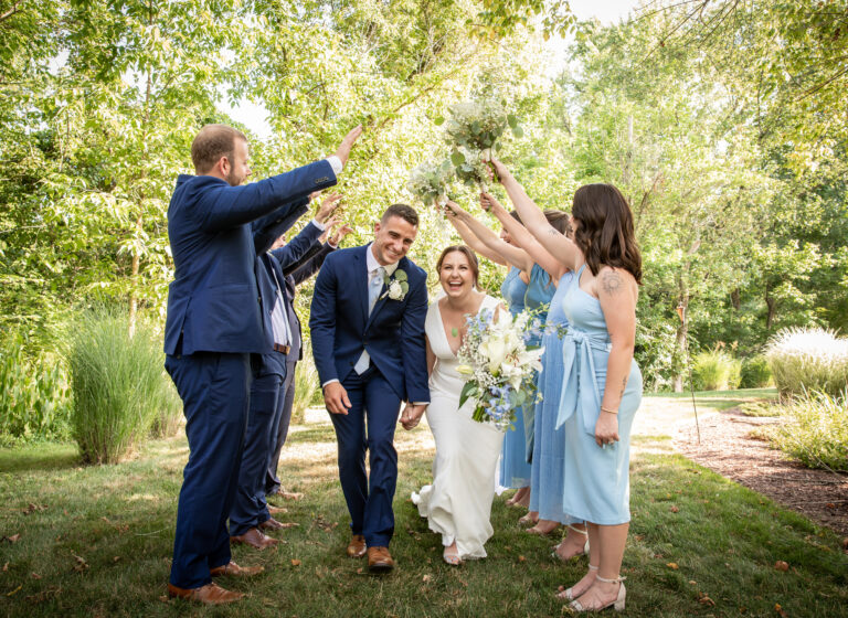 Bride and groom walk under raised arms of wedding party outdoors, surrounded by greenery.