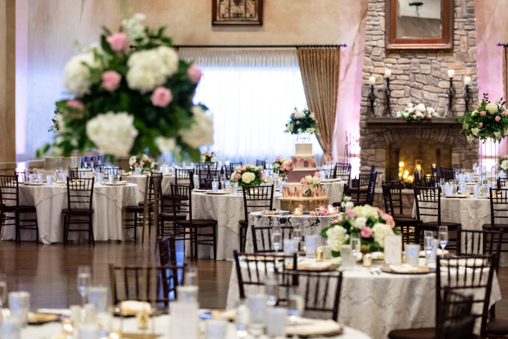 Elegant wedding reception with floral centerpieces, a tiered cake, round tables, and a lit stone fireplace.