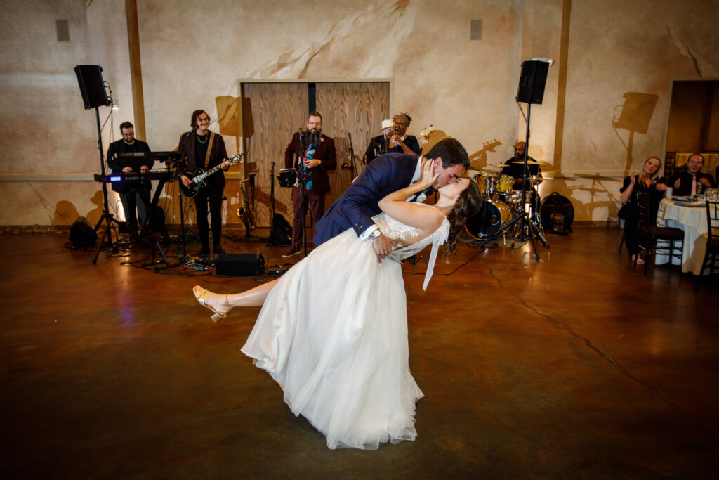 A newlywed couple shares a kiss on the dance floor, with a band playing in the background.