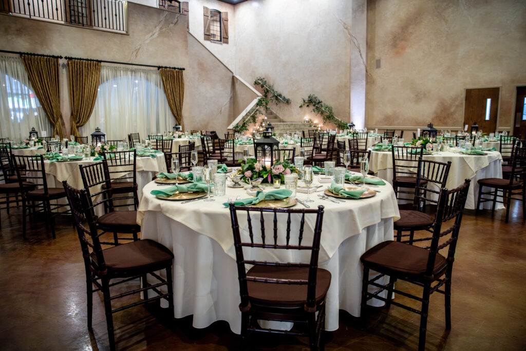 Elegant banquet hall with round tables, white tablecloths, green napkins, and dark wooden chairs; staircase in the background.