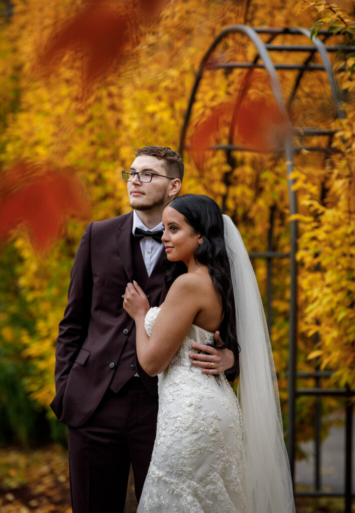 Bride and groom standing under autumn leaves, the groom in a suit and the bride in a lace dress and veil.