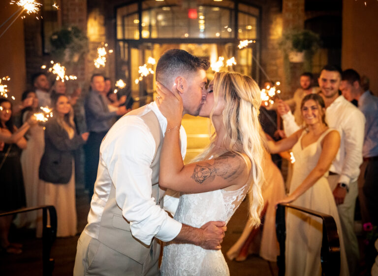 A couple kisses at a wedding, surrounded by guests holding sparklers.
