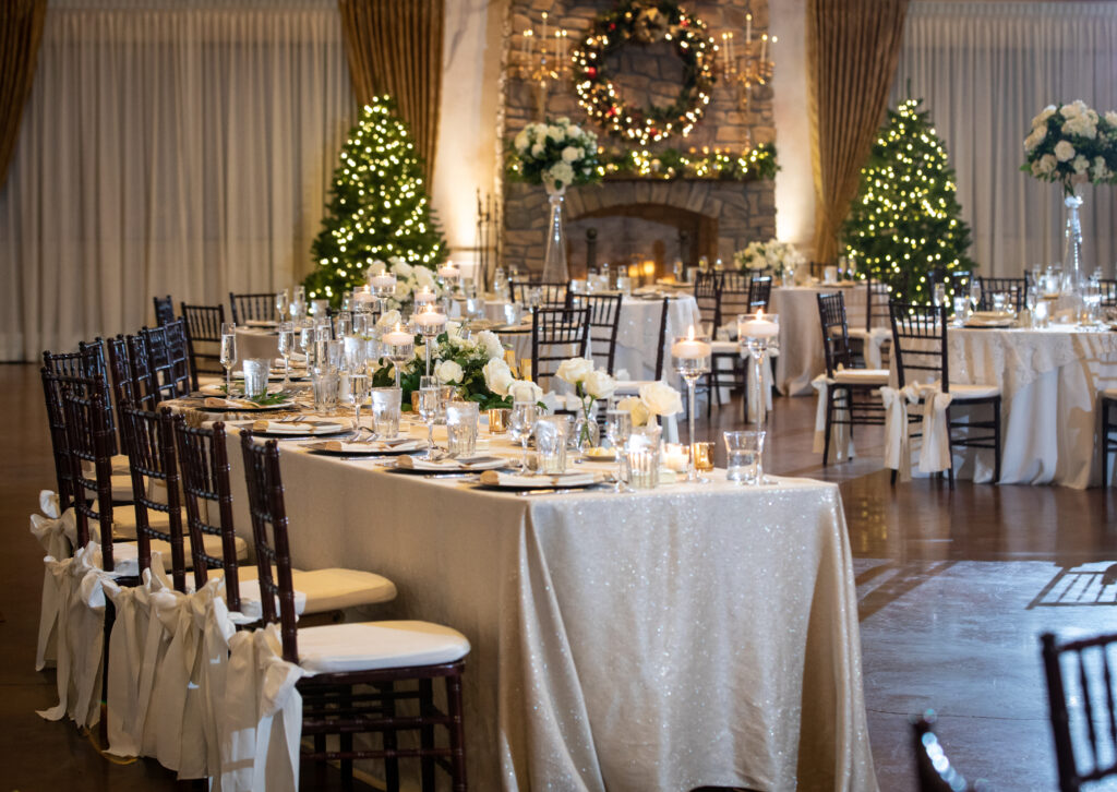 Elegant banquet hall with decorated tables, white flowers, candles, Christmas wreath, and trees with lights.