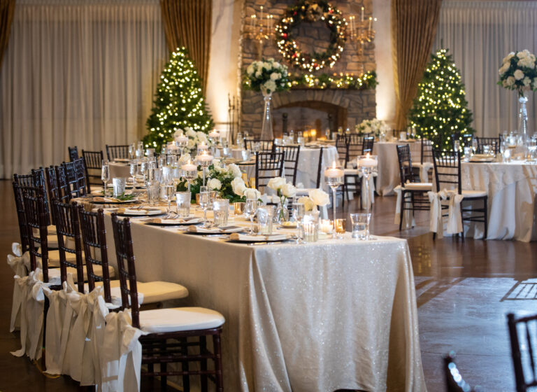 Elegant banquet hall with decorated tables, white flowers, candles, Christmas wreath, and trees with lights.