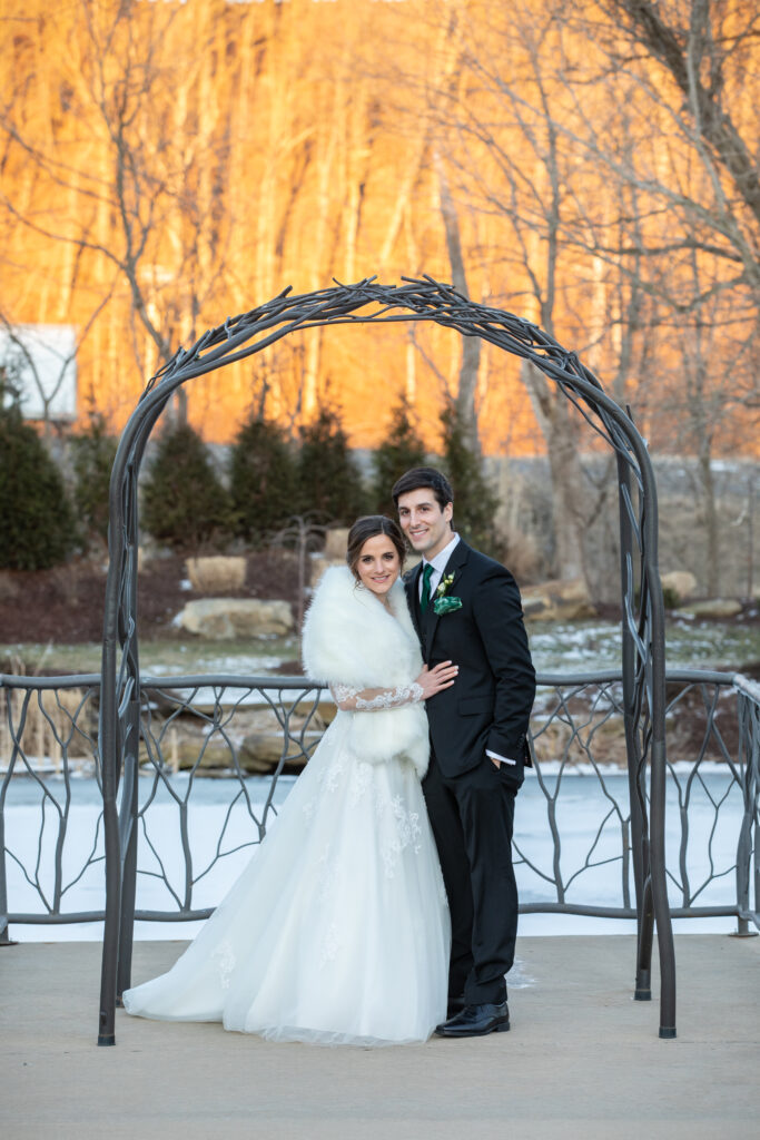 Bride and groom standing under a metal arch outdoors, with trees in the background.