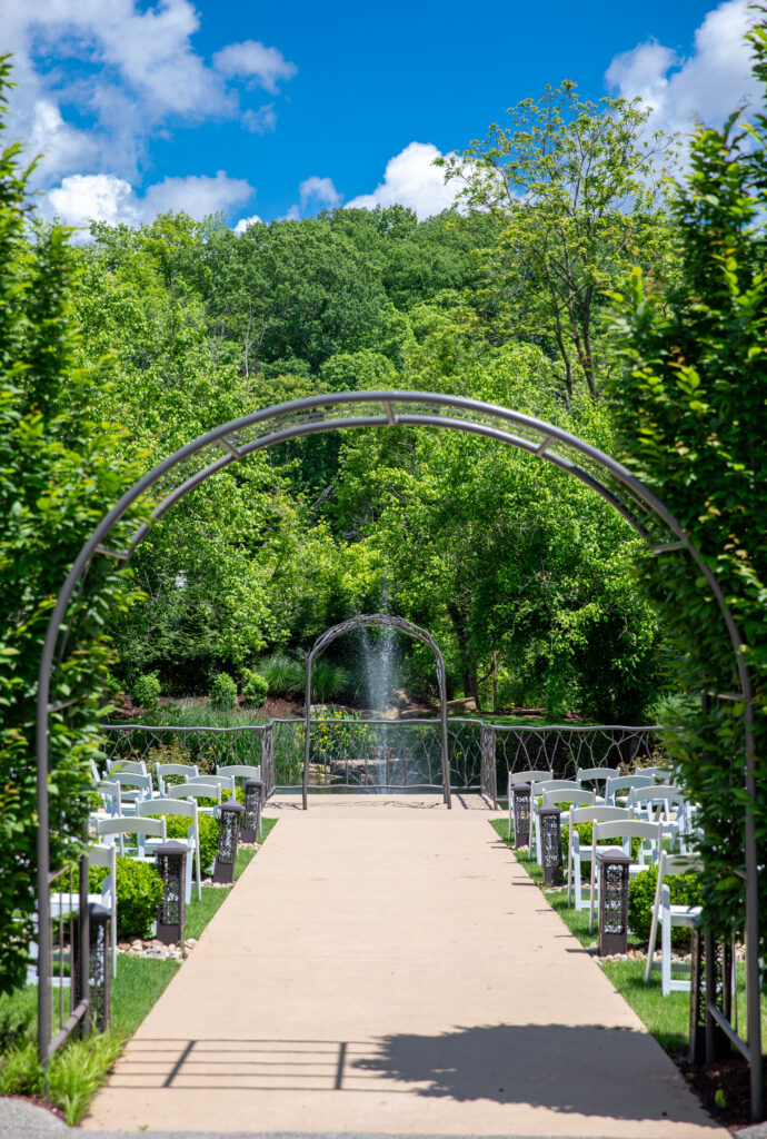 Outdoor wedding aisle with white chairs, metal arches, and a fountain, surrounded by lush green trees and blue sky.