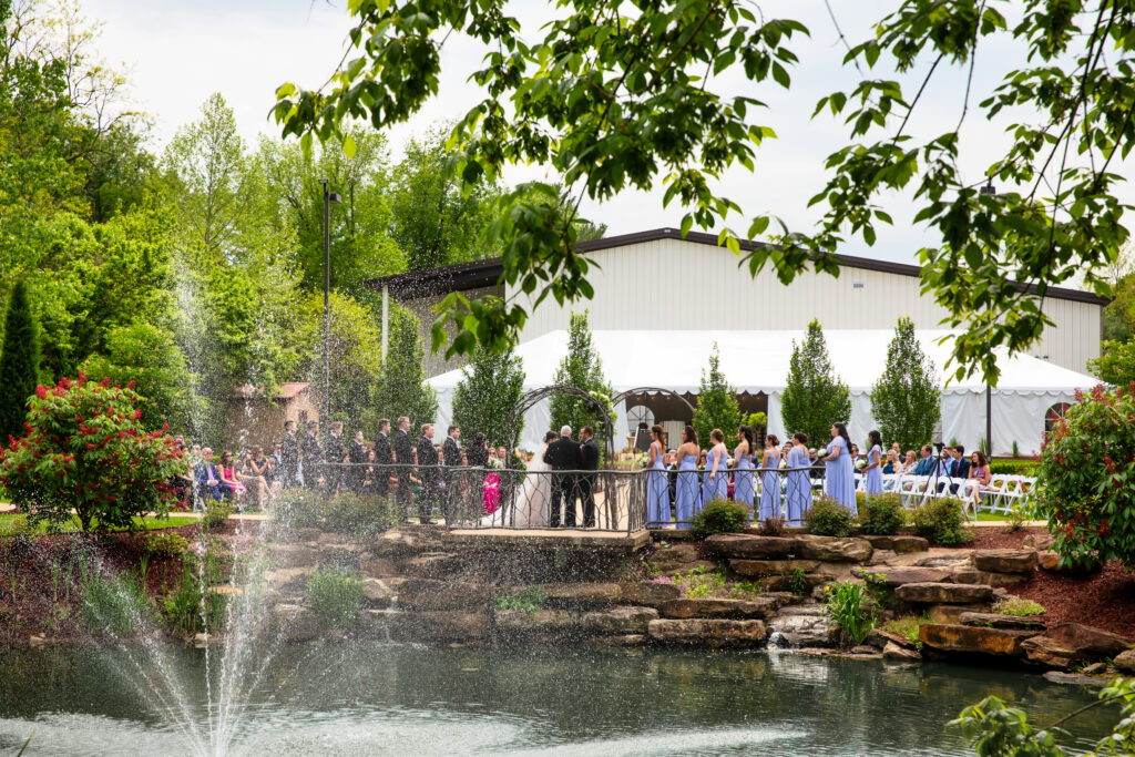 Outdoor wedding ceremony by a pond with a fountain, surrounded by greenery and attended by guests under a white tent.