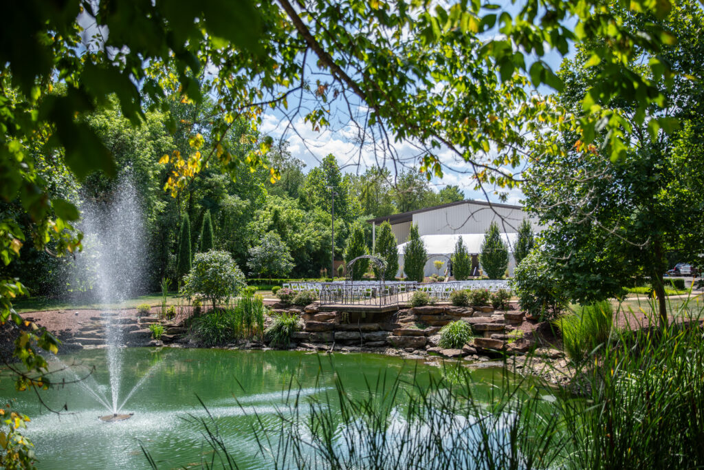 A serene garden pond with a water fountain, surrounded by lush greenery and a white building in the background.