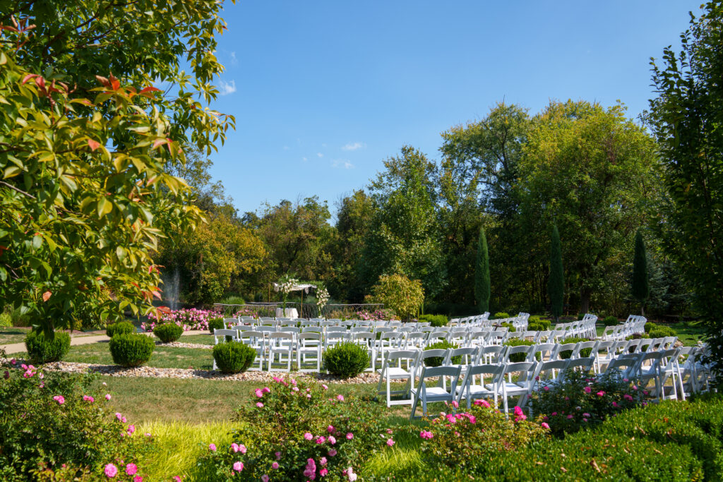 Outdoor wedding setup with rows of white chairs and pink flowers, surrounded by lush green trees and bushes.