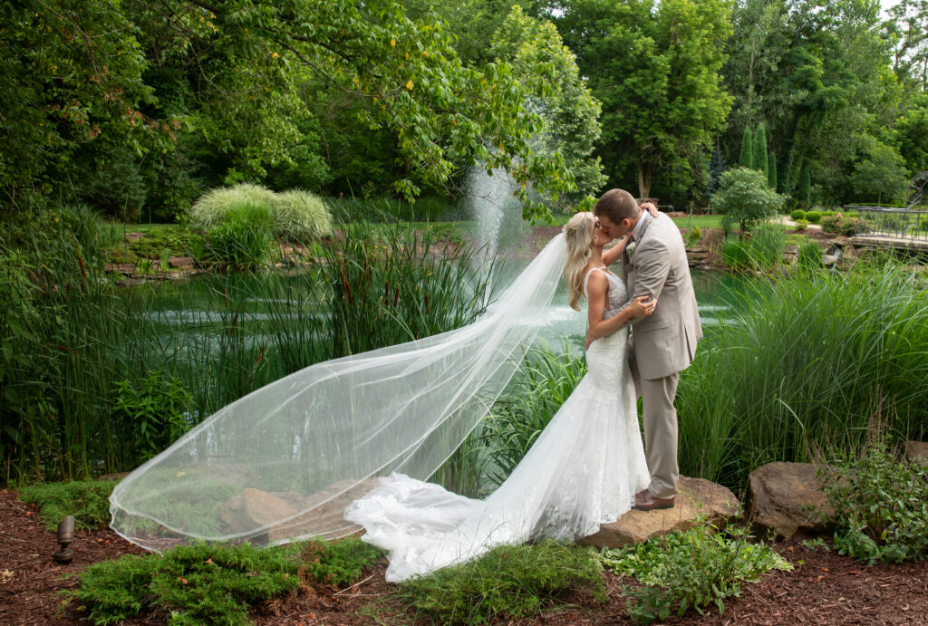A couple kisses by a pond, surrounded by greenery. The bride's veil flows gracefully in the breeze.