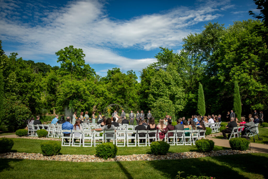 Outdoor wedding ceremony with guests seated in white chairs, surrounded by green trees under a partly cloudy sky.