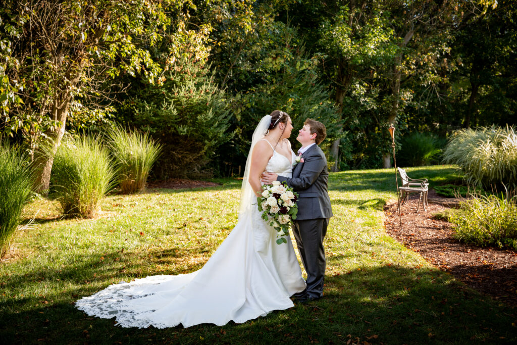 A bride and groom stand in a garden, embracing and smiling at each other, with greenery and a bench in the background.