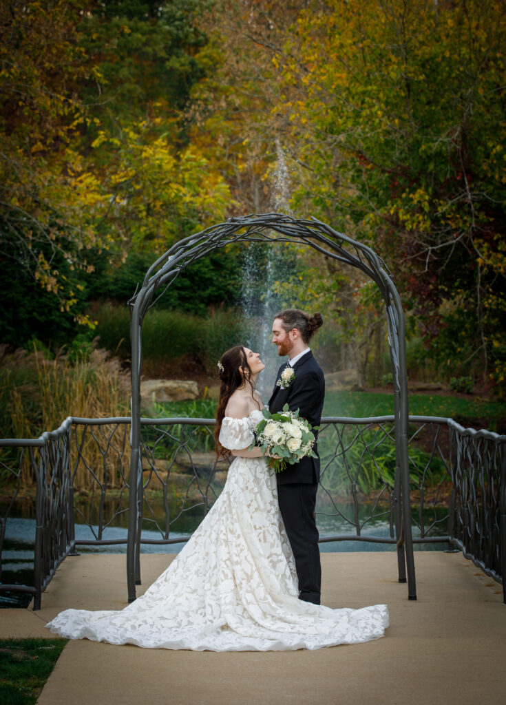 A couple in wedding attire embrace under an archway with a fountain and autumn foliage in the background.