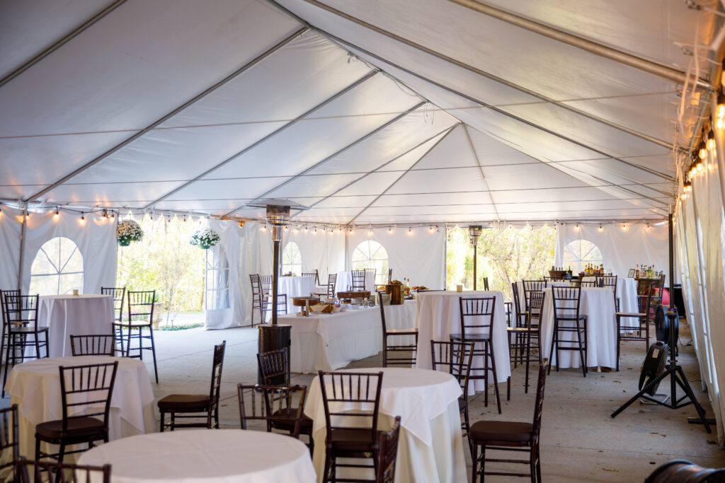 Interior of a decorated event tent with round tables, chairs, string lights, and a buffet table.