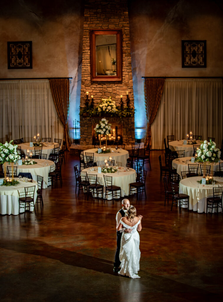 A couple dances alone in an elegant, dimly lit hall with decorated round tables and a large stone fireplace.