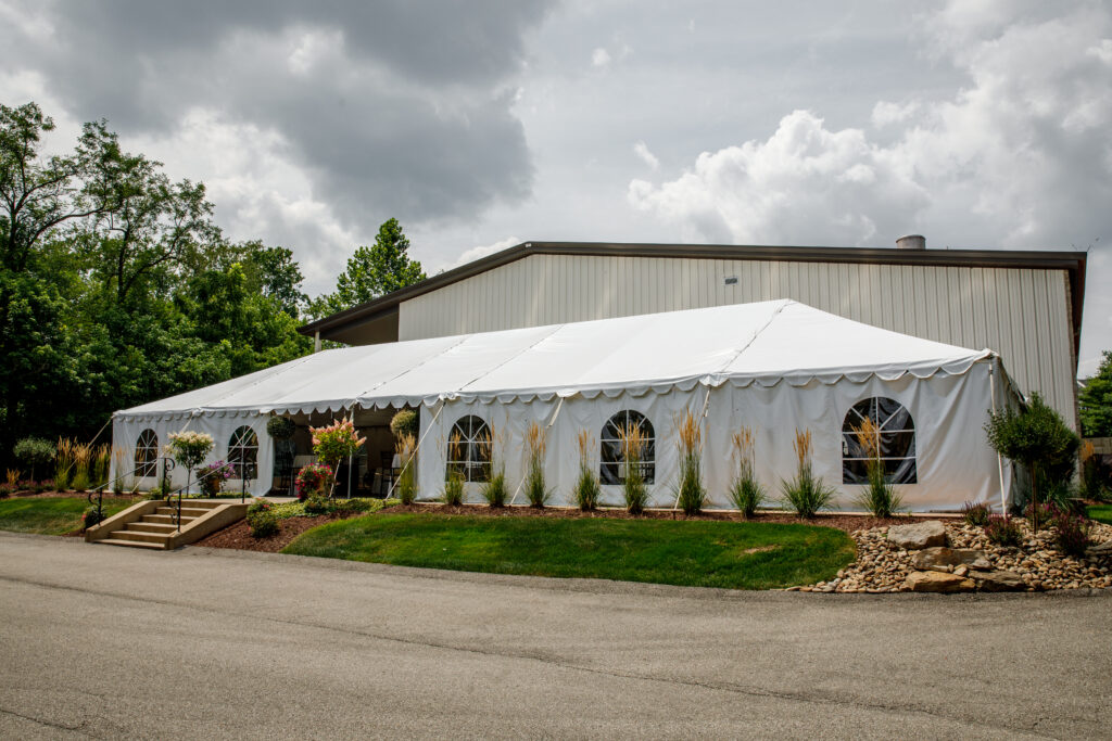 Large white event tent adjacent to a building, surrounded by greenery and ornamental plants. Cloudy sky above.