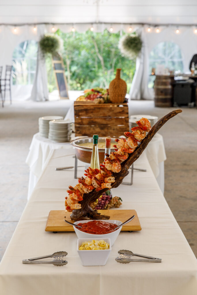 Skewered shrimp on a wooden stand with a bowl of sauce, on a table in a tented event setting.