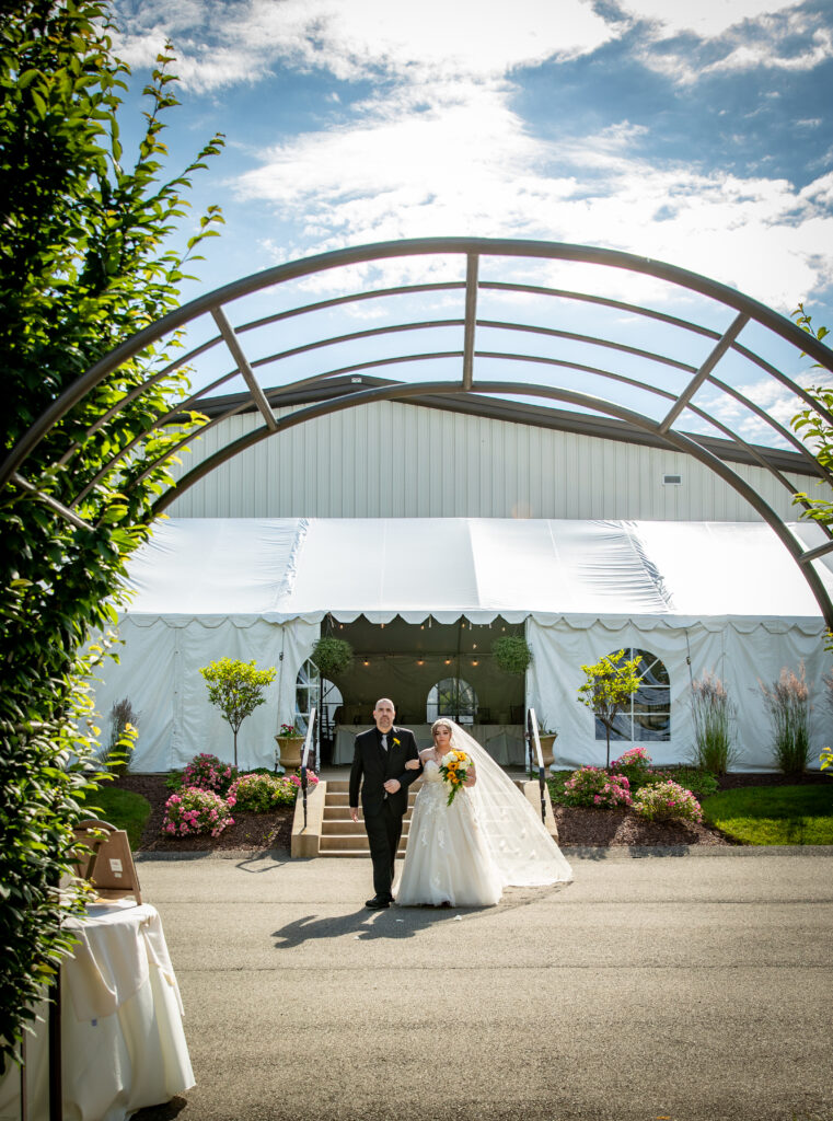 Bride and groom walking under a metal archway towards a white tent on a sunny day, surrounded by flowers.