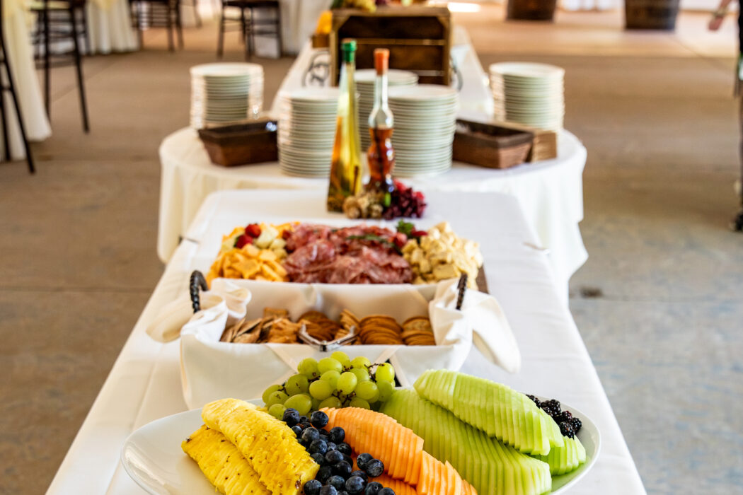 Buffet table with assorted fruits, meats, cheeses, and plates set in a sunlit, elegant event tent.