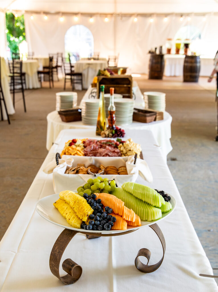 Buffet table with assorted fruits, meats, cheeses, and plates set in a sunlit, elegant event tent.