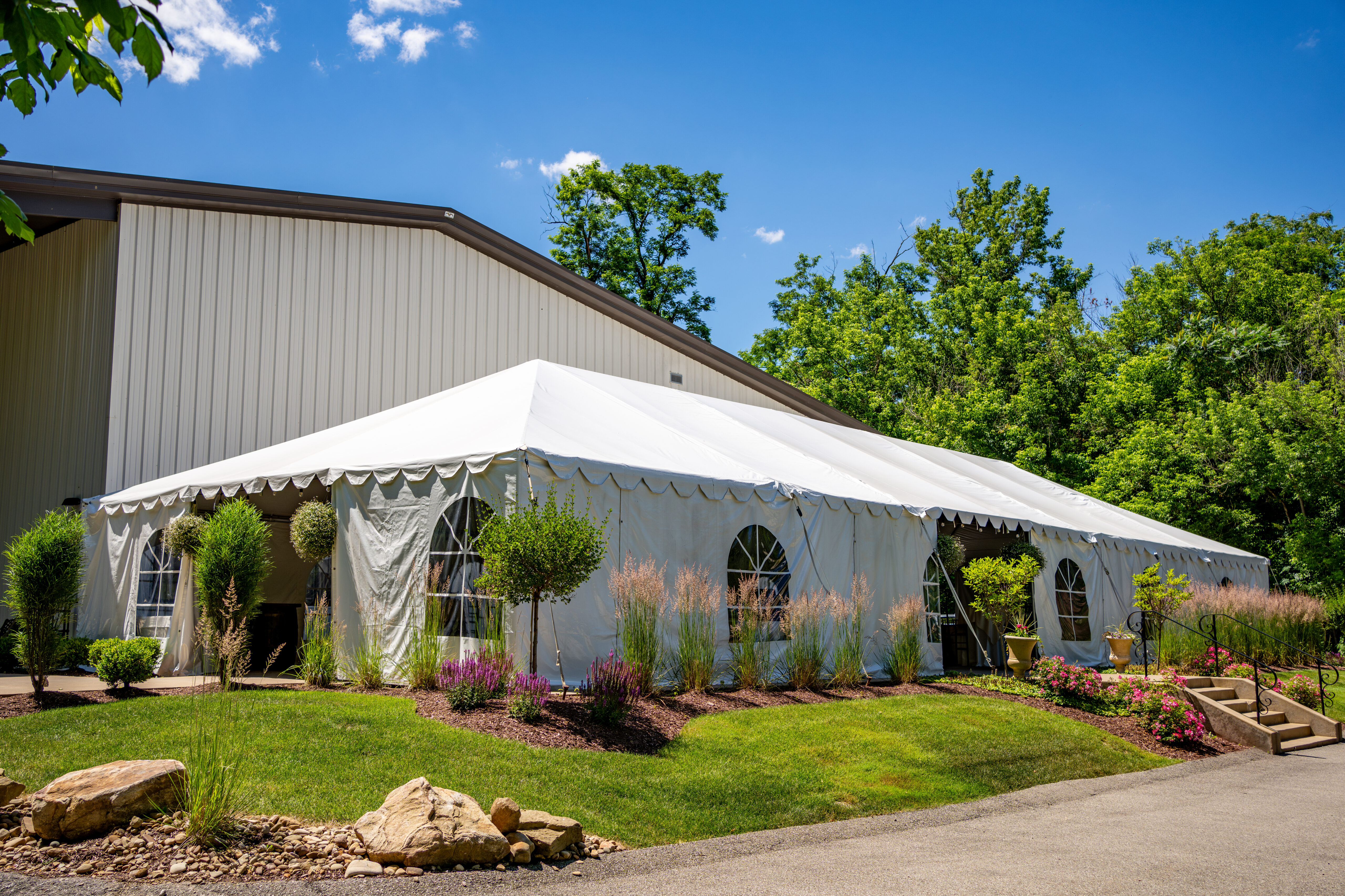 A white event tent with arched windows stands near a building, surrounded by greenery and clear blue skies.