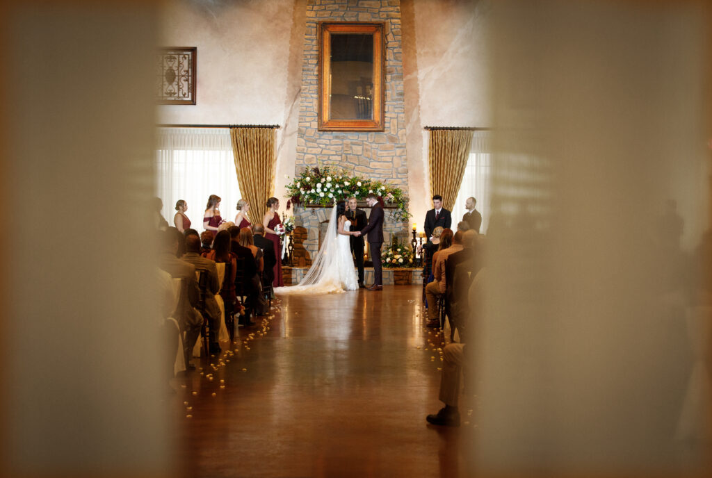 A couple stands at the altar during a wedding ceremony, framed by curtains, in a hall with guests seated on both sides.