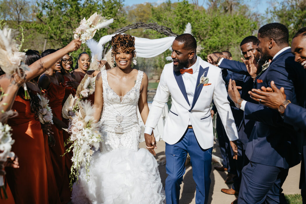 Bride and groom joyfully walk down the aisle, surrounded by cheering guests with greenery in the background.