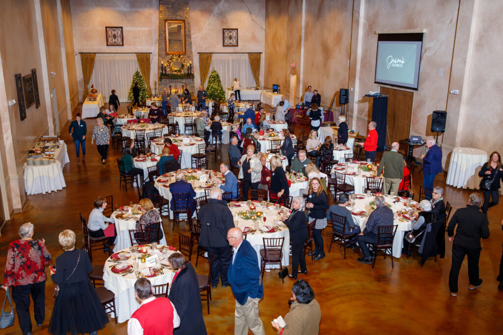 A large dining hall filled with people sitting at round tables, decorated with Christmas trees and a projector screen.