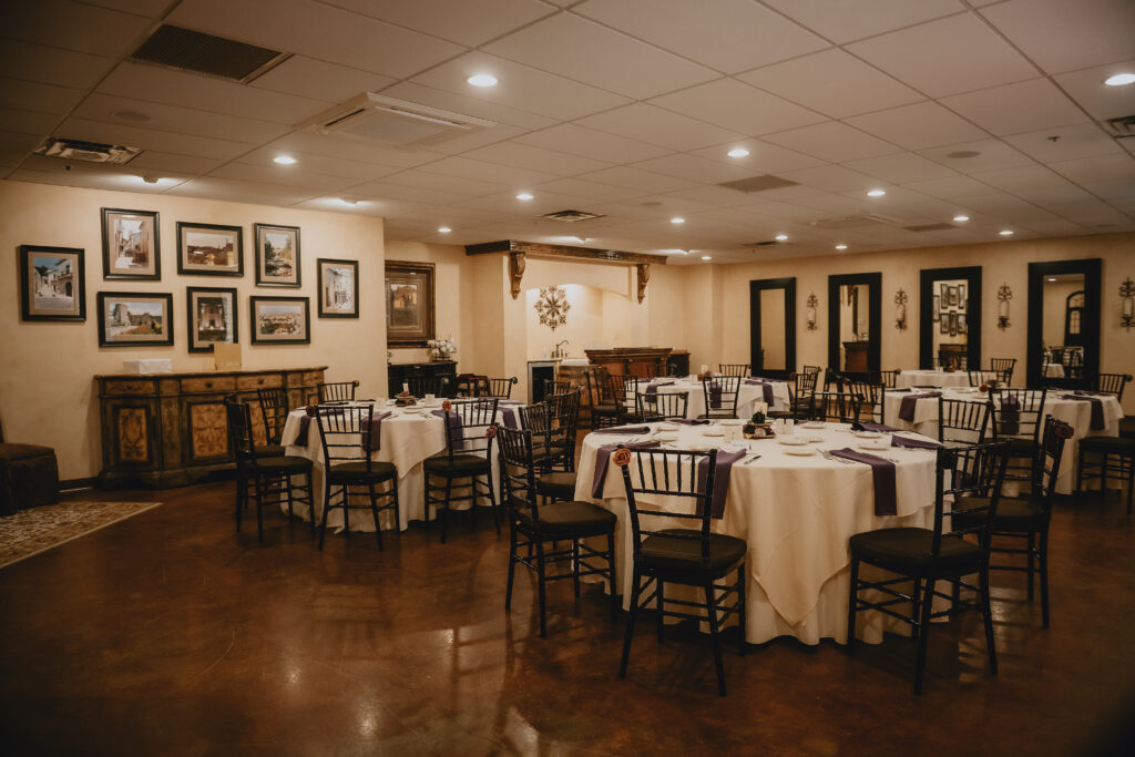 A banquet hall with round tables, white tablecloths, black chairs, and framed pictures on beige walls.