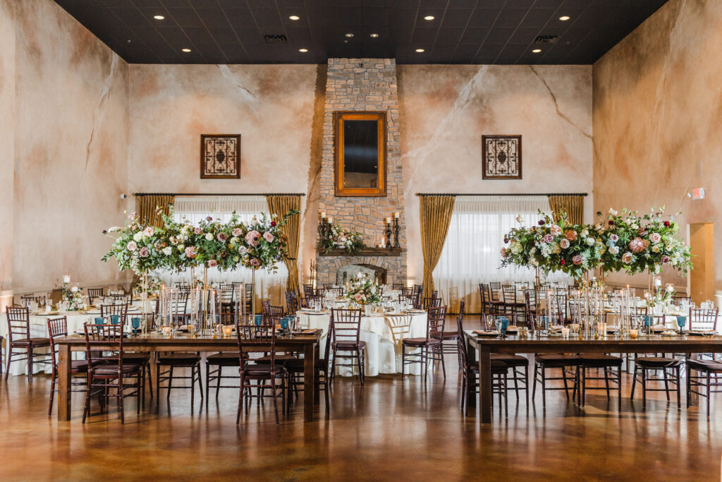 Elegant banquet hall with long tables, tall floral centerpieces, wooden chairs, and a rustic stone fireplace.