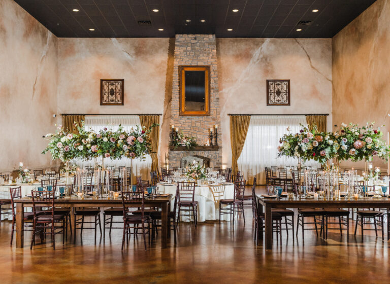 Elegant banquet hall with long tables, tall floral centerpieces, wooden chairs, and a rustic stone fireplace.