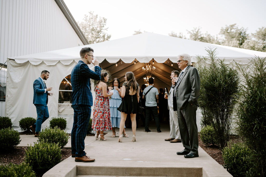 People socialize outside a white tent at an outdoor event, with plants and a chandelier visible inside.