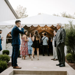 People socialize outside a white tent at an outdoor event, with plants and a chandelier visible inside.