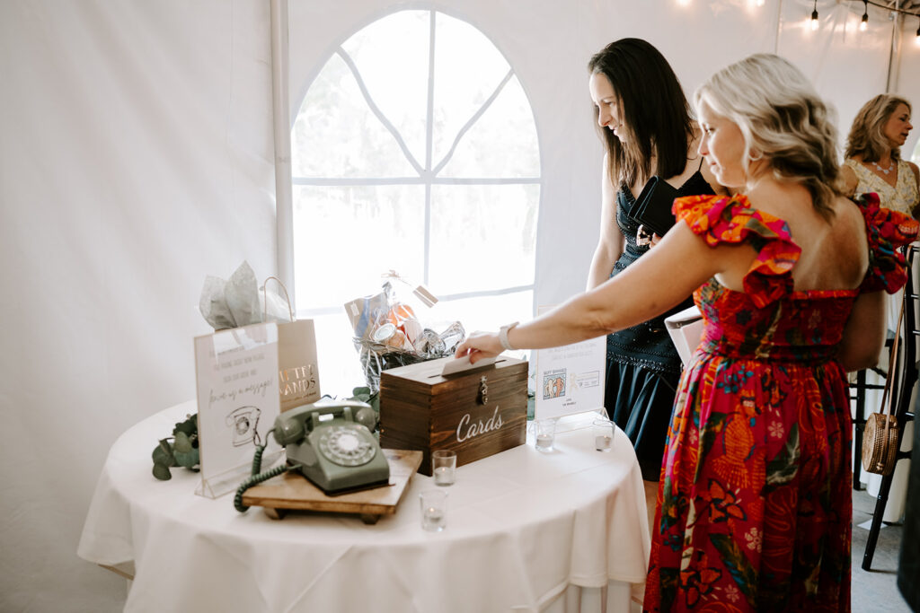 Two women stand at a table with a card box, gifts, and a vintage phone at an event.