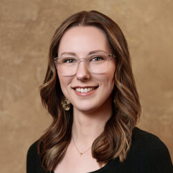 Person with long brown hair, glasses, and dimple smiling, wearing a dark top and gold earrings against a beige background.