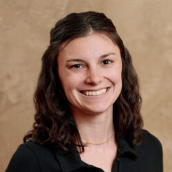 Smiling woman with wavy brown hair wearing a black shirt against a beige background.