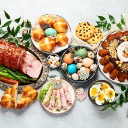 A festive table with ham, asparagus, decorated eggs, bread, pastries, cookies, and deviled eggs, garnished with leaves.