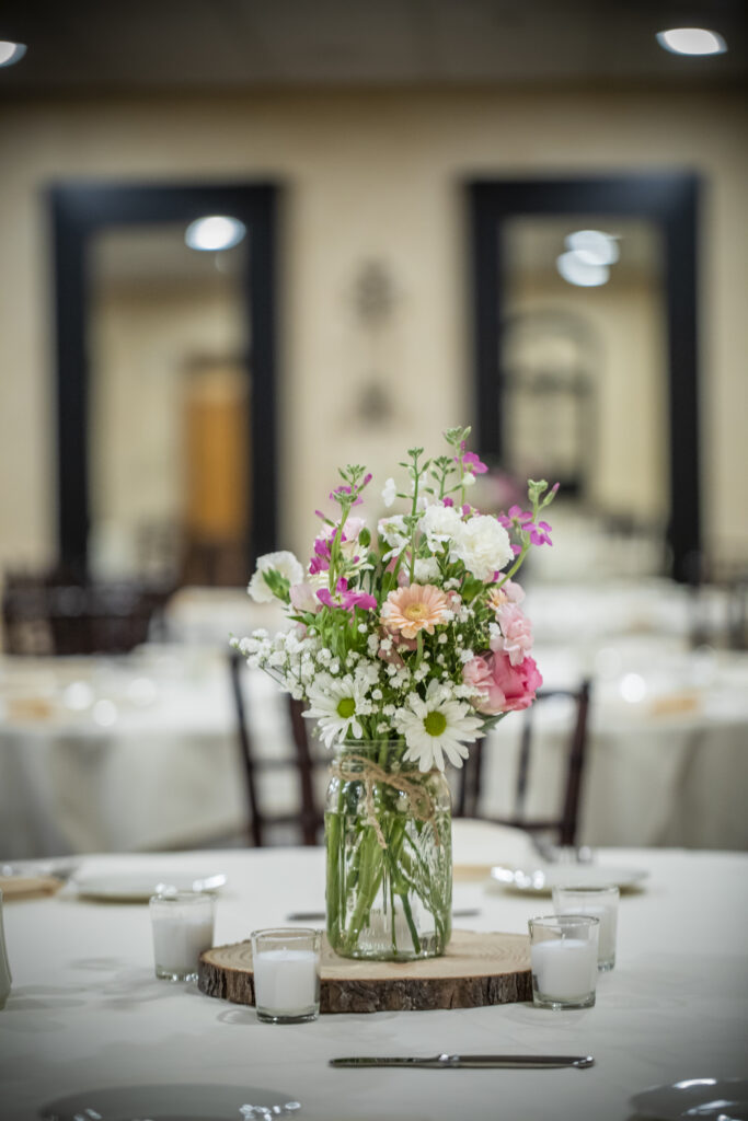 Floral arrangement in a glass jar on a wooden slice, surrounded by candles, on a table in a banquet setting.